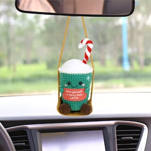 Könnte beinhalten: Eine grüne und weiße gehäkelte Kaffeetasse mit einem roten und weißen Zuckerrohr, das am Rand hängt. Die Tasse hat ein lächelndes Gesicht und den Text "Have yourself a merry little LATTE!" auf der Vorderseite. Die Tasse hängt an einem Rückspiegel in einem Auto.