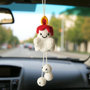 Op de afbeelding: Een handgemaakte, witte gehaakte kaars auto-hanger met een lachend gezicht, rode was en een gele vlam. Twee witte gehaakte ballen met lachende gezichten hangen eronder. De hanger hangt aan een wit touwtje.