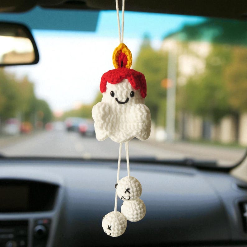 Op de afbeelding: Een handgemaakte, witte gehaakte kaars auto-hanger met een lachend gezicht, rode was en een gele vlam. Twee witte gehaakte ballen met lachende gezichten hangen eronder. De hanger hangt aan een wit touwtje.