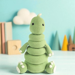 Op de afbeelding: Een groene gehaakte stapeltoren in de vorm van een rups. Het speelgoed heeft een afgeronde kop, een gesegmenteerd lichaam en kleine armen en voeten met witte accenten. De achtergrond is een lichtblauwe muur met decoratieve elementen.