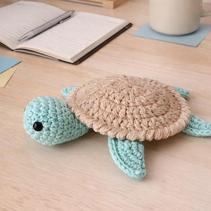 May include: A handmade crochet turtle in shades of light blue and tan. The turtle has a textured shell, head, and flippers. The turtle's head has a black eye. The turtle is lying on a wooden surface.