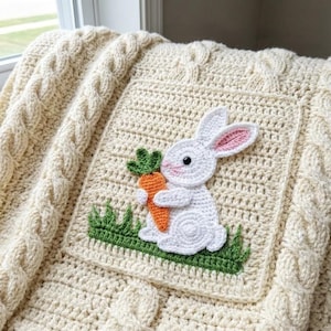 Pode incluir: Cobertor de croch&ecirc; creme com borda de malha de cabo. Um patch quadrado apresenta um coelho branco de croch&ecirc; segurando uma cenoura laranja, sobre grama verde. O coelho tem orelhas internas rosa e um olho preto.