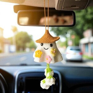 Op de afbeelding: Een handgemaakte, witte gehaakte auto-hanger in de vorm van een spook, met een bruine hoed en een lachend gezicht. Het heeft gele en roze bloemaccenten en een tros witte bloemen die eronder hangen. De hanger hangt aan de achteruitkijkspiegel van een auto.
