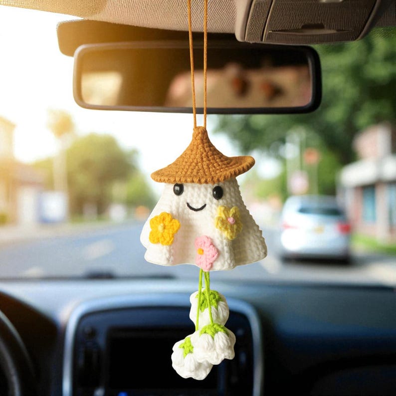 Op de afbeelding: Een handgemaakte, witte gehaakte auto-hanger in de vorm van een spook, met een bruine hoed en een lachend gezicht. Het heeft gele en roze bloemaccenten en een tros witte bloemen die eronder hangen. De hanger hangt aan de achteruitkijkspiegel van een auto.