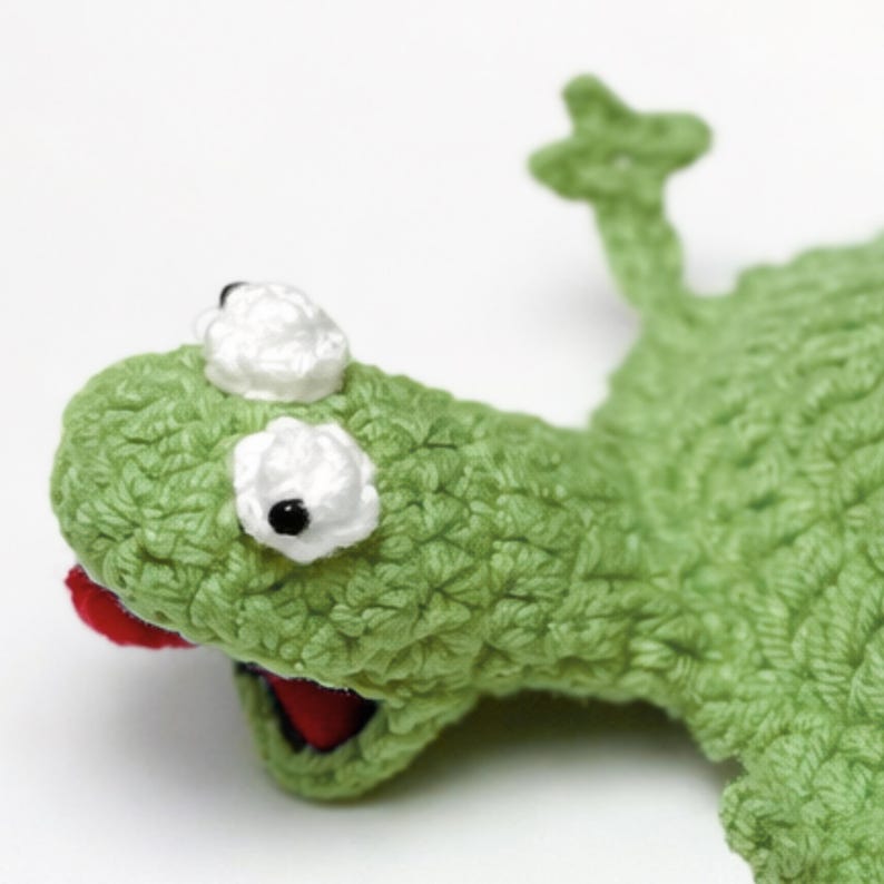 Puede incluir: Un lagarto verde hecho a mano con ojos blancos y boca roja. El lagarto est&aacute; tejido con hilo texturizado, lo que le da una apariencia tridimensional. El dise&ntilde;o incluye una peque&ntilde;a cabeza levantada y una cola.