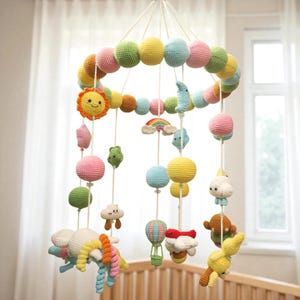 May include: A colorful, handmade baby mobile featuring a ring of crocheted spheres in pastel colors. Hanging below are crocheted suns, clouds, rainbows, and other whimsical shapes. The mobile is suspended from a white string, designed to hang over a crib.