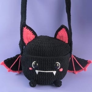 May include: A black crochet bat-shaped crossbody bag with pink detailing. The bag has large, round eyes, a white mouth with two fangs, and pink-lined ears and wings. The strap is black.