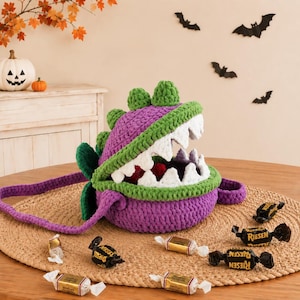 May include: A purple and green crocheted monster bag with a wide-open mouth featuring white teeth. The bag has a long purple strap and is surrounded by several individually wrapped chocolate candies on a round, textured, woven surface.