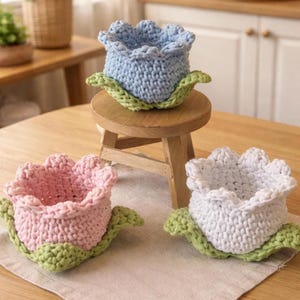 Puede incluir: Tres cestas de flores de ganchillo hechas a mano en colores pastel: azul claro, rosa y blanco. Cada cesta tiene un borde festoneado y descansa sobre una base de hoja verde. Las cestas se exhiben sobre una mesa de madera.