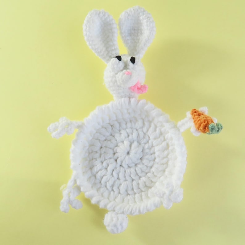 May include: A handmade white crochet bunny with long ears, black eyes, and a pink mouth. The bunny holds a small orange carrot with green leaves. The bunny is on a yellow background.