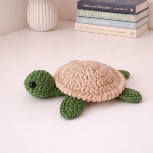 May include: A handmade crochet turtle toy. The turtle has a light tan shell, green head, and green flippers. The turtle is sitting on a white surface. Books are in the background.