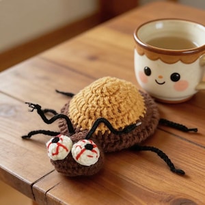 May include: A handmade crochet spider coaster with a brown body, tan back, and black legs. The spider has white eyes with red veins. A ceramic mug with a smiling face is in the background.