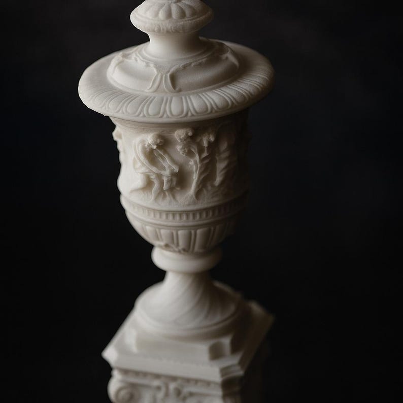 Pu&ograve; includere: Un'urna bianca e ornata con un'intagliatura a rilievo dettagliata di figure e fogliame. L'urna ha una base a piedistallo e un coperchio a cupola. Il design &egrave; classico, con un focus sulla simmetria e sulla consistenza. L'estetica generale &egrave; antica ed elegante.