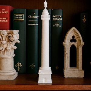 Puede incluir: Una réplica blanca en miniatura de una columna alta con una estatua en la parte superior, de pie sobre una estantería de madera. La columna está flanqueada por libros de tapa dura verdes y elementos arquitectónicos decorativos de piedra. Los libros tienen letras doradas.