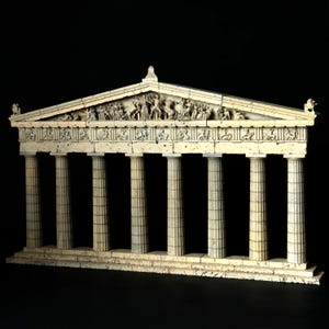 May include: A cream-colored architectural model of a classical Greek temple featuring eight fluted columns, a detailed pediment with sculpted figures, and a frieze depicting various scenes. The structure has a weathered, stone-like texture.