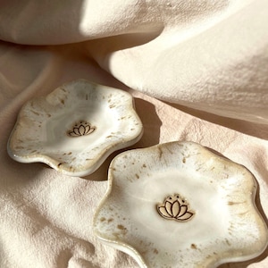 Könnte beinhalten: Zwei Keramikschalen mit weiß-beiger Glasur und einem Lotusblüten-Design. Die Schalen haben eine organische, wellenförmige Form. Die Lotusblüte ist dunkelbraun. Die Schalen liegen auf einem hellrosa Stoff.