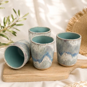May include: A set of four ceramic tumblers featuring a reactive glaze with shades of turquoise, blue, and white. The cups have a smooth, cylindrical shape with a solid turquoise interior and a textured, mottled exterior finish in cool tones.