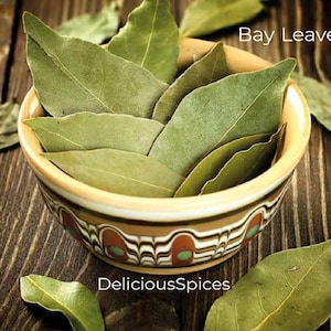 May include: A bowl filled with dried bay leaves, a common culinary herb. The bowl is decorated with a colorful pattern and the text "DeliciousSpices" is visible below the bowl. The image is set against a wooden background.