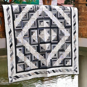 May include: A black and white quilt with a log cabin pattern. The quilt is hanging on a railing over a body of water. The quilt has a white border with a black and white braided design.