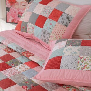 Puede incluir: Una colcha rosa de patchwork con patrones rojos, grises y florales. La colcha est&aacute; hecha de cuadrados de diferentes telas cosidos juntos. La colcha est&aacute; en una cama con dos almohadas a juego.