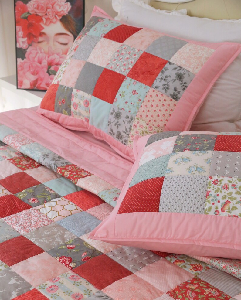 Puede incluir: Una colcha rosa de patchwork con patrones rojos, grises y florales. La colcha est&aacute; hecha de cuadrados de diferentes telas cosidos juntos. La colcha est&aacute; en una cama con dos almohadas a juego.