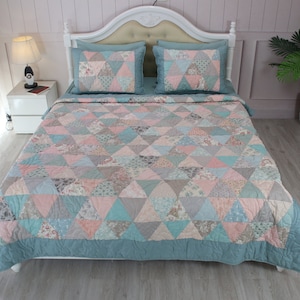 Puede incluir: Una colcha azul turquesa con un diseño de patchwork de triángulos rosas, grises y blancos. La colcha está sobre una cama con dos fundas de almohada a juego.