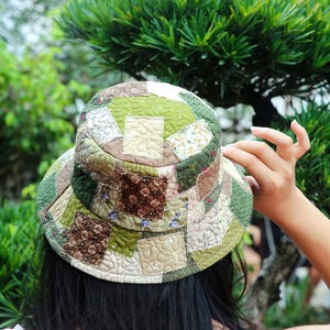 May include: A patchwork hat in green, brown, and cream. The hat has a wide brim and a rounded crown, with various fabric squares sewn together. The design includes floral and geometric patterns, creating a unique, handcrafted look. The hat is a stylish accessory.