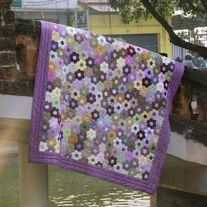 Puede incluir: Un edredón hecho a mano con un estampado floral en tonos morados, verdes, amarillos y marrones. El edredón presenta un diseño de flores hexagonales y un borde morado liso. El edredón está drapeado sobre una estructura de piedra.