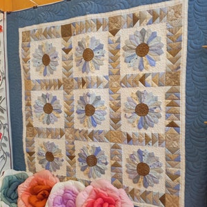 以下が含まれることがあります： 青と茶色の花柄のキルト。キルトは、それぞれ異なるデザインの小さな正方形で構成されています。キルトは壁に掛けられています。