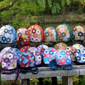 Peut inclure: Une collection de 15 sacs à dos en patchwork colorés fabriqués à partir de chutes de tissu. Les sacs à dos sont disposés sur un banc en pierre dans un jardin. Chaque sac à dos présente un design unique de formes de fleurs hexagonales.