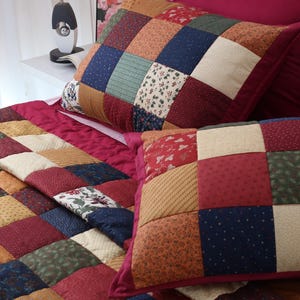 May include: A patchwork quilt and two matching pillow shams featuring a grid of square fabric patches in various patterns and colors, including deep red, navy blue, forest green, mustard yellow, and cream with floral and geometric prints.