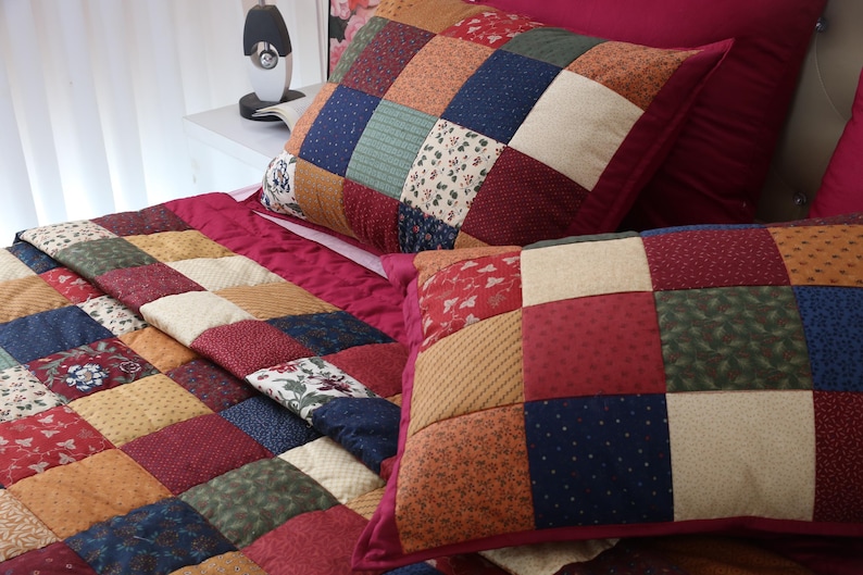 May include: A patchwork quilt and two matching pillow shams featuring a grid of square fabric patches in various patterns and colors, including deep red, navy blue, forest green, mustard yellow, and cream with floral and geometric prints.