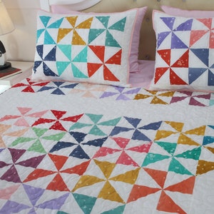 May include: A white quilt with a colorful pinwheel pattern. The quilt is made of various shades of blue, green, red, pink, and yellow. The quilt is on a bed with two matching pillowcases.
