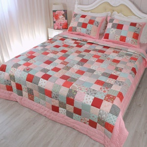 Puede incluir: Un edred&oacute;n de retazos rosa y gris con un patr&oacute;n floral. El edred&oacute;n est&aacute; hecho de cuadrados de diferentes telas y colores. El edred&oacute;n est&aacute; en una cama con almohadas blancas.
