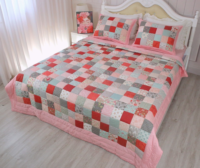 Puede incluir: Un edred&oacute;n de retazos rosa y gris con un patr&oacute;n floral. El edred&oacute;n est&aacute; hecho de cuadrados de diferentes telas y colores. El edred&oacute;n est&aacute; en una cama con almohadas blancas.