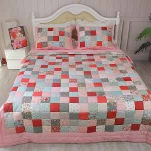 May include: A patchwork quilt with a pink, red and grey colour scheme. The quilt is made of squares of different fabrics, each with a floral pattern. The quilt is on a bed with two matching pillowcases.