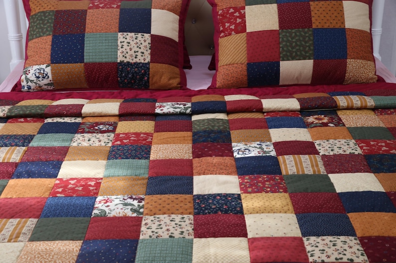 May include: A patchwork quilt and two matching pillow shams featuring a grid of square fabric patches in various colors including deep red, navy blue, forest green, mustard yellow, and cream, with assorted floral and geometric patterns.