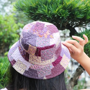 以下が含まれることがあります： 紫、ラベンダー、バーガンディの色合いのキルトパッチワークバケットハット。帽子は、幅広いブリムと丸みを帯びたクラウンを備え、さまざまな花柄と幾何学模様が特徴です。帽子は布製で、日焼け防止用に設計されています。