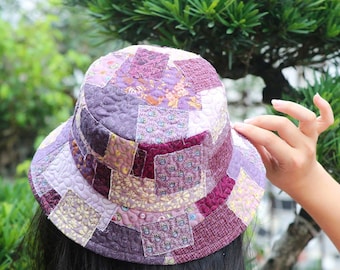 May include: A quilted patchwork bucket hat in shades of purple, lavender, and burgundy. The hat features a variety of floral and geometric patterns, with a wide brim and a rounded crown. The hat is made of fabric and is designed for sun protection.