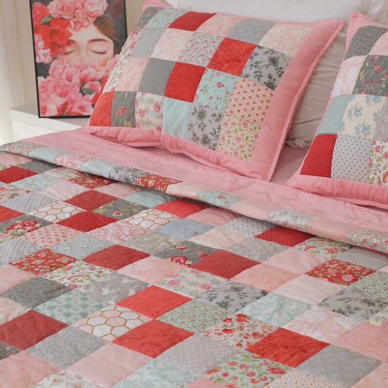 Puede incluir: Una colcha de patchwork rosa con una variedad de patrones florales y geom&eacute;tricos en tonos de rosa, rojo, gris y azul. La colcha est&aacute; hecha de cuadrados y est&aacute; doblada sobre una cama con dos almohadas de patchwork a juego.