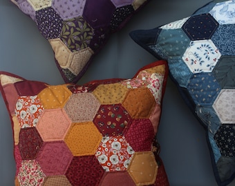 Peut inclure: Trois coussins en patchwork avec des motifs hexagonaux. Les coussins sont faits de différents tissus avec des motifs floraux et géométriques. Les coussins sont dans des tons de violet, bleu, rouge, orange et jaune.