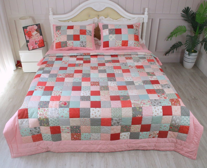 Puede incluir: Un edred&oacute;n de patchwork con un borde rosa y una variedad de patrones florales y geom&eacute;tricos en tonos de rojo, rosa, gris y verde. El edred&oacute;n est&aacute; sobre una cama blanca con dos fundas de almohada a juego.