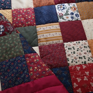 May include: A patchwork quilt featuring a variety of square fabric patches in shades of burgundy, navy blue, forest green, mustard yellow, and cream. The squares display diverse patterns including floral prints, small dots, stripes, and leaf motifs.