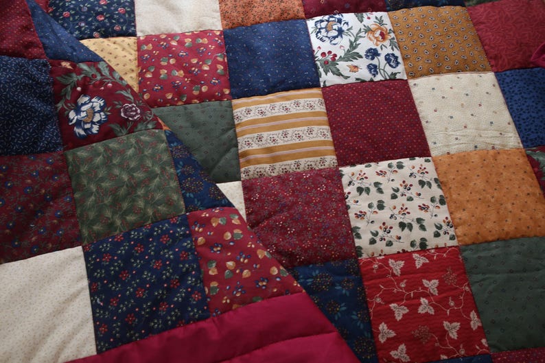 May include: A patchwork quilt featuring a variety of square fabric patches in shades of burgundy, navy blue, forest green, mustard yellow, and cream. The squares display diverse patterns including floral prints, small dots, stripes, and leaf motifs.