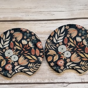 May include: Two black round cup coasters with a floral pattern. The pattern features beige, orange, and white flowers and leaves on a black background.