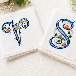 Pode incluir: Dois lenços brancos bordados com as letras "V" e "S" em azul e dourado. As letras são decoradas com abóboras laranja e folhas verdes. As bordas dos lenços são finalizadas com renda delicada.