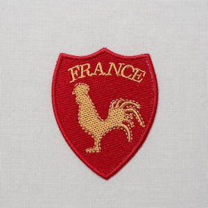 Peut inclure: Écusson rouge brodé avec un coq doré et le mot "FRANCE" en or. Le patch en forme de bouclier est un symbole de la France, possiblement pour une équipe sportive ou la fierté nationale.