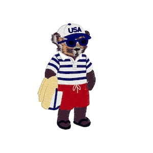 Pode incluir: Urso bordado vestindo uma camisa polo listrada em branco e azul, shorts vermelhos, óculos de sol e um boné de beisebol dos EUA. O urso está segurando uma toalha de praia e um pequeno livro azul e branco. O urso está usando sandálias pretas.