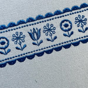 May include: Close-up of a blue embroidered floral design on a light gray fabric. The design features stylized flowers and scalloped edges, creating a decorative border. The embroidery is detailed and the color contrast is striking.