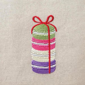 May include: An embroidered design on beige fabric featuring a vertical stack of four colorful macarons in green, pink, cream, and purple. A bright red embroidered ribbon is tied around the entire stack of macarons, forming a bow at the top.